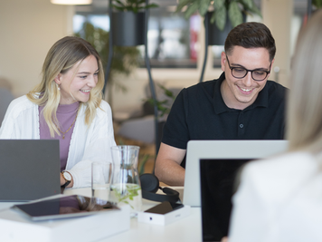 Eine Mitarbeiterin und ein Mitarbeiter lächeln und arbeiten an Laptops, während sie an der Highbench sitzen, auf dem ein Tablet und ein Glas Wasser stehen. Im Hintergrund befindet sich der Cafébereich.
