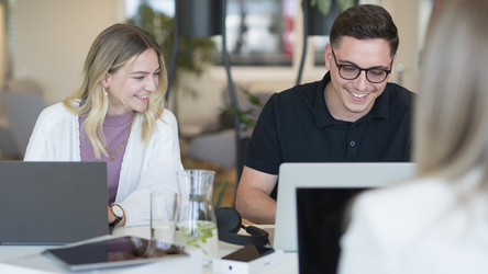 Eine Mitarbeiterin und ein Mitarbeiter lächeln und arbeiten an Laptops, während sie an der Highbench sitzen, auf dem ein Tablet und ein Glas Wasser stehen. Im Hintergrund befindet sich der Cafébereich.