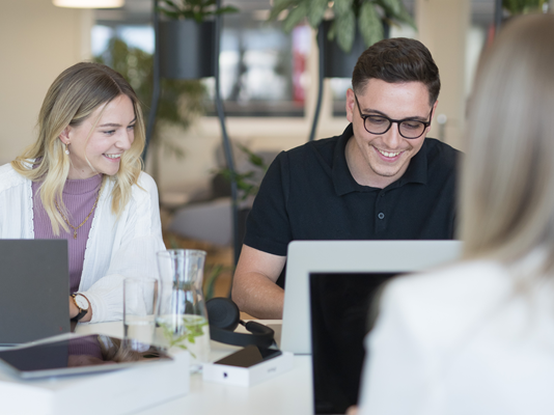 Eine Mitarbeiterin und ein Mitarbeiter lächeln und arbeiten an Laptops, während sie an der Highbench sitzen, auf dem ein Tablet und ein Glas Wasser stehen. Im Hintergrund befindet sich der Cafébereich.