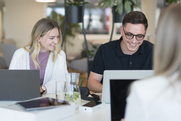 Eine Mitarbeiterin und ein Mitarbeiter lächeln und arbeiten an Laptops, während sie an der Highbench sitzen, auf dem ein Tablet und ein Glas Wasser stehen. Im Hintergrund befindet sich der Cafébereich.