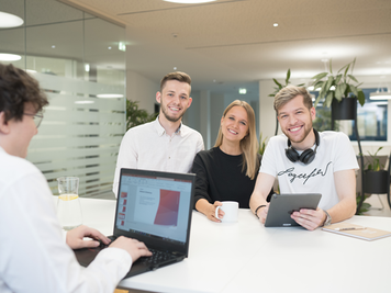 Vier Mitarbeitende in einem Büro, von denen drei in die Kamera lächeln. Ein Mitarbeiter arbeitet am Laptop, während ein anderer ein Tablet hält. Im Hintergrund sind Pflanzen zu sehen.