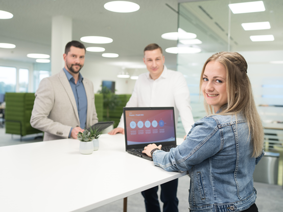 Zwei Mitarbeiter und eine Mitarbeiterin stehen gemeinsam um eine Highbench im Büro. Die Mitarbeiterin hat den Laptop aufgeklappt, und alle lächeln in die Kamera.