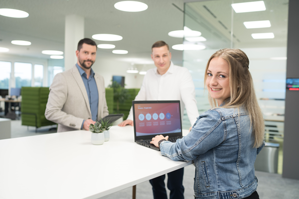 Zwei Mitarbeiter und eine Mitarbeiterin stehen gemeinsam um eine Highbench im Büro. Die Mitarbeiterin hat den Laptop aufgeklappt, und alle lächeln in die Kamera.