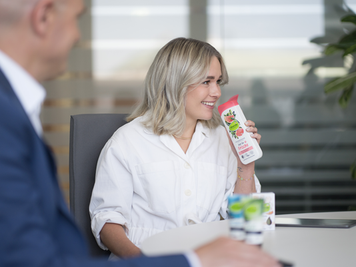 Eine Mitarbeiterin sitzt am Besprechungstisch und riecht an einem dm Shampoo. Vor ihr stehen weitere Produkte, und im Vordergrund ist die Silhouette eines Mitarbeiters zu sehen.