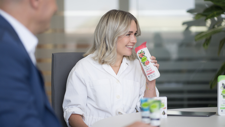Eine Mitarbeiterin sitzt am Besprechungstisch und riecht an einem dm Shampoo. Vor ihr stehen weitere Produkte, und im Vordergrund ist die Silhouette eines Mitarbeiters zu sehen.