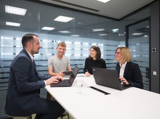 Zwei Mitarbeiter und zwei Mitarbeiterinnen sitzen gemeinsam in einem Besprechungsraum an der Highbench. Sie diskutieren lächelnd miteinander und haben Laptops und Tablets vor sich.