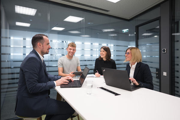 Zwei Mitarbeiter und zwei Mitarbeiterinnen sitzen gemeinsam in einem Besprechungsraum an der Highbench. Sie diskutieren lächelnd miteinander und haben Laptops und Tablets vor sich.