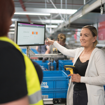 Im Lager erklärt eine Mitarbeiterin einer Kollegin etwas, während sie auf einen Bildschirm zeigt, der Informationen anzeigt. In der Hand hält sie ein Handscanner-Gerät. Im Hintergrund sind blaue Lagerkisten und weitere Mitarbeitende sichtbar, die beschäftigt sind.