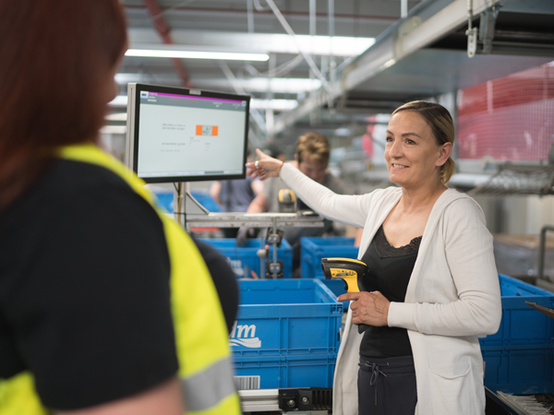 Im Lager erklärt eine Mitarbeiterin einer Kollegin etwas, während sie auf einen Bildschirm zeigt, der Informationen anzeigt. In der Hand hält sie ein Handscanner-Gerät. Im Hintergrund sind blaue Lagerkisten und weitere Mitarbeitende sichtbar, die beschäftigt sind.