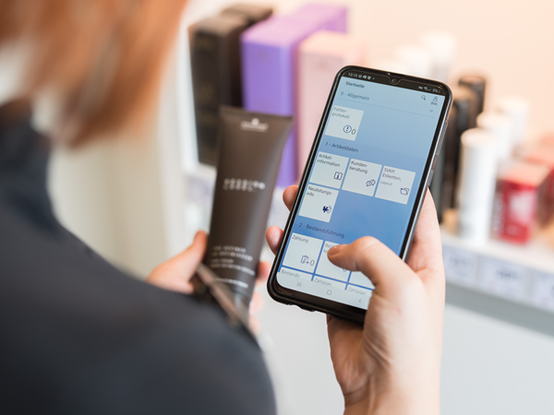 Eine Person steht vor einem Regal in einem dm Markt und hält ein Smartphone in der einen und ein Produkt in der anderen Hand. Auf dem Smartphone ist eine App geöffnet, die Informationen zu Produkten anzeigt.