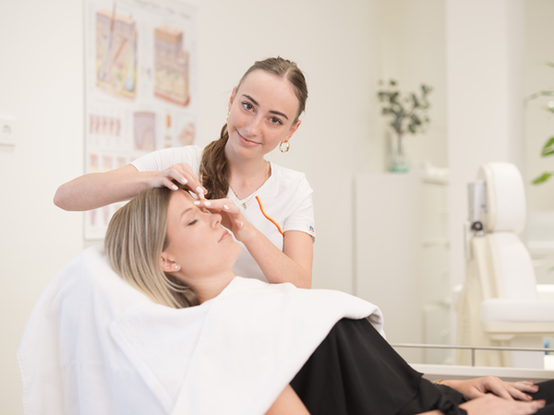 Eine Mitarbeiterin führt eine  Gesichtsbehandlung durch, während die Kundin mit geschlossenen Augen entspannt auf einem Behandlungsstuhl. Im Hintergrund sind informative Poster und Pflanzen sichtbar, die zur angenehmen Umgebung beitragen.