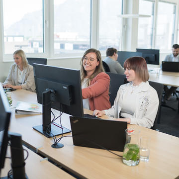 Mitarbeitend  arbeitet in einem modernen Büro. Eine Person zeigt etwas auf dem Bildschirm einer Kollegin, während diese aufmerksam zuschaut. Im Hintergrund sind weitere Mitarbeiter an ihren Schreibtischen zu sehen, und die Umgebung ist hell und einladend, mit großen Fenstern, die Tageslicht hereinlassen. Auf dem Tisch stehen Pflanzen und Unterlagen, die zur angenehmen Arbeitsatmosphäre beitragen.