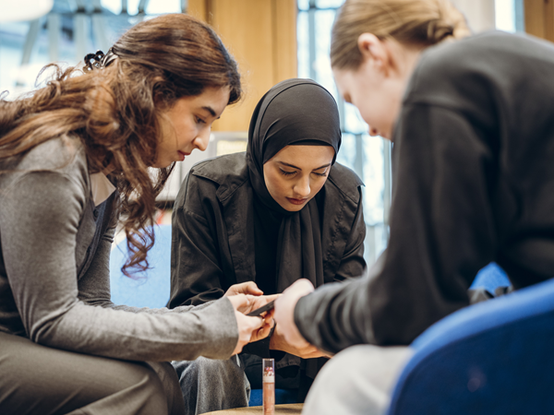Eine Gruppe von Lehrlingen befindet sich bei einem Workshop.  Sie schauen auf ein Gerät oder eine Vorlage und Atmosphäre ist konzentriert und kooperativ.