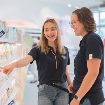 Eine Mitarbeiterin in einem dm Markt zeigt auf ein Produkt im Regal, während sie mit einer Kollegin spricht. Beide wirken engagiert und interessiert an den Produkten, die sie besprechen.
