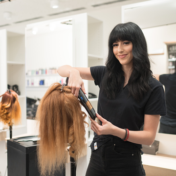 Eine Lehrling in einer dm Akademie verwendet ein Stylinggerät, um die Haare eines Übungskopfes zu stylen. Im Hintergrund ist ein weiterer Lehrling zu sehen, der ebenfalls mit einer Puppe arbeitet. Die Akademie ist modern eingerichtet und mit verschiedenen Haarpflegeprodukten ausgestattet.