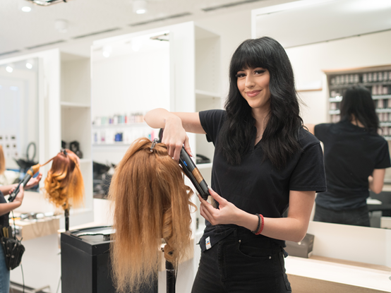 Eine Lehrling in einer dm Akademie verwendet ein Stylinggerät, um die Haare eines Übungskopfes zu stylen. Im Hintergrund ist ein weiterer Lehrling zu sehen, der ebenfalls mit einer Puppe arbeitet. Die Akademie ist modern eingerichtet und mit verschiedenen Haarpflegeprodukten ausgestattet.