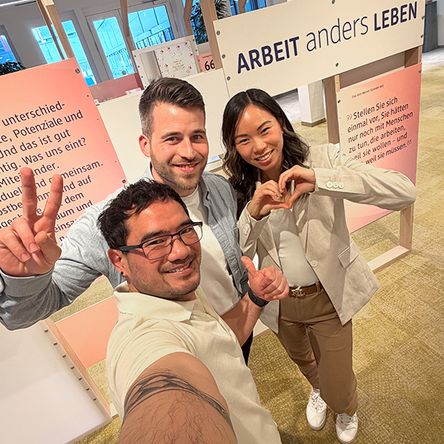 Drei lächelnde Mitarbeitende posieren für ein Selfie in einem Eingansbereich in einem Büro. Hinter ihnen ist ein Schild mit der Aufschrift Arbeit anders leben zu sehen.