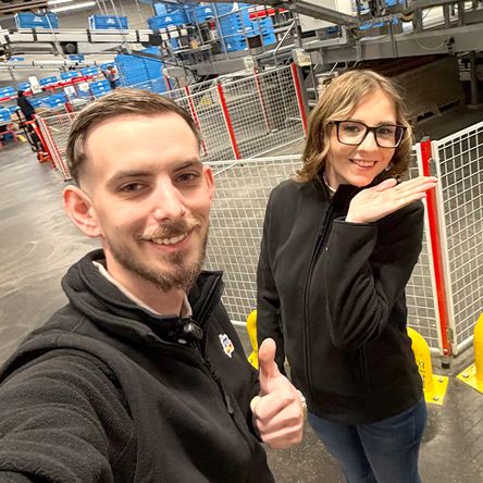 Zwei Mitarbeitende befinden sich im Lagerbereich im Verteilzentrum. Die beiden machen ein Selfie von sich und ein Mitarbeiter macht einen Daumen nach oben.