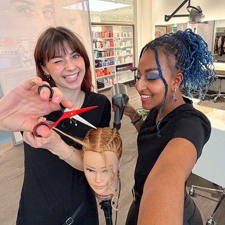 Zwei Mitarbeitende sind in einem Friseurstudio und üben an einer Puppe das Haareschneiden. Die Puppe hat eine coole Frisur. Die eine Kollegin hält spielerisch eine Schere in der Hand und zwinkert, während die andere einen Föhn benutzt. Beide lächeln und die Athmosphäre ist locker.