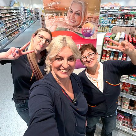 Drei Mitarbeitende in Arbeitskleidung posieren in einem dm Markt vor einem bunten Plakat. Die Kolleginnen haben Spaß und zwei davon machen ein Peace-Zeichen. Die Athmosphäre wirkt modern und hell.