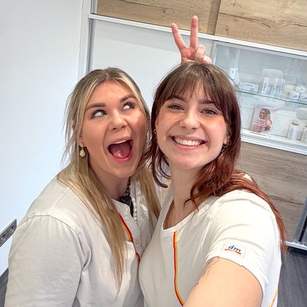 Zwei Mitarbeiterinnen befinden sich in einem modernen Kosmetikstudio und machen ein Selfie von sich. Die Athmosphäre wirkt sehr locker und die beiden haben Spaß. Eine Kollegin macht ein Peace Zeichen hinter dem Kopf von der anderen Kollegin.