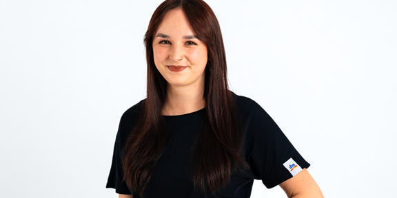 Ein weiblicher Lehrling, die vor einem neutralen Hintergrund steht und in die Kamera lächelt. Eine Hand hat sie in die Hüfte gestützt.