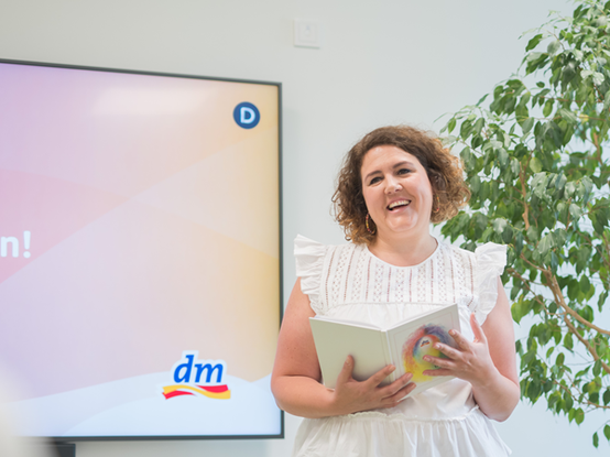 Eine Frau in einer weißen Bluse lächelt und hält das dm Leitbild in der Hand, während sie neben einem Bildschirm steht, der das "dm"-Logo zeigt.