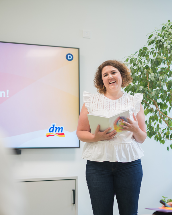 Eine Frau in einer weißen Bluse lächelt und hält das dm Leitbild in der Hand, während sie neben einem Bildschirm steht, der das "dm"-Logo zeigt.