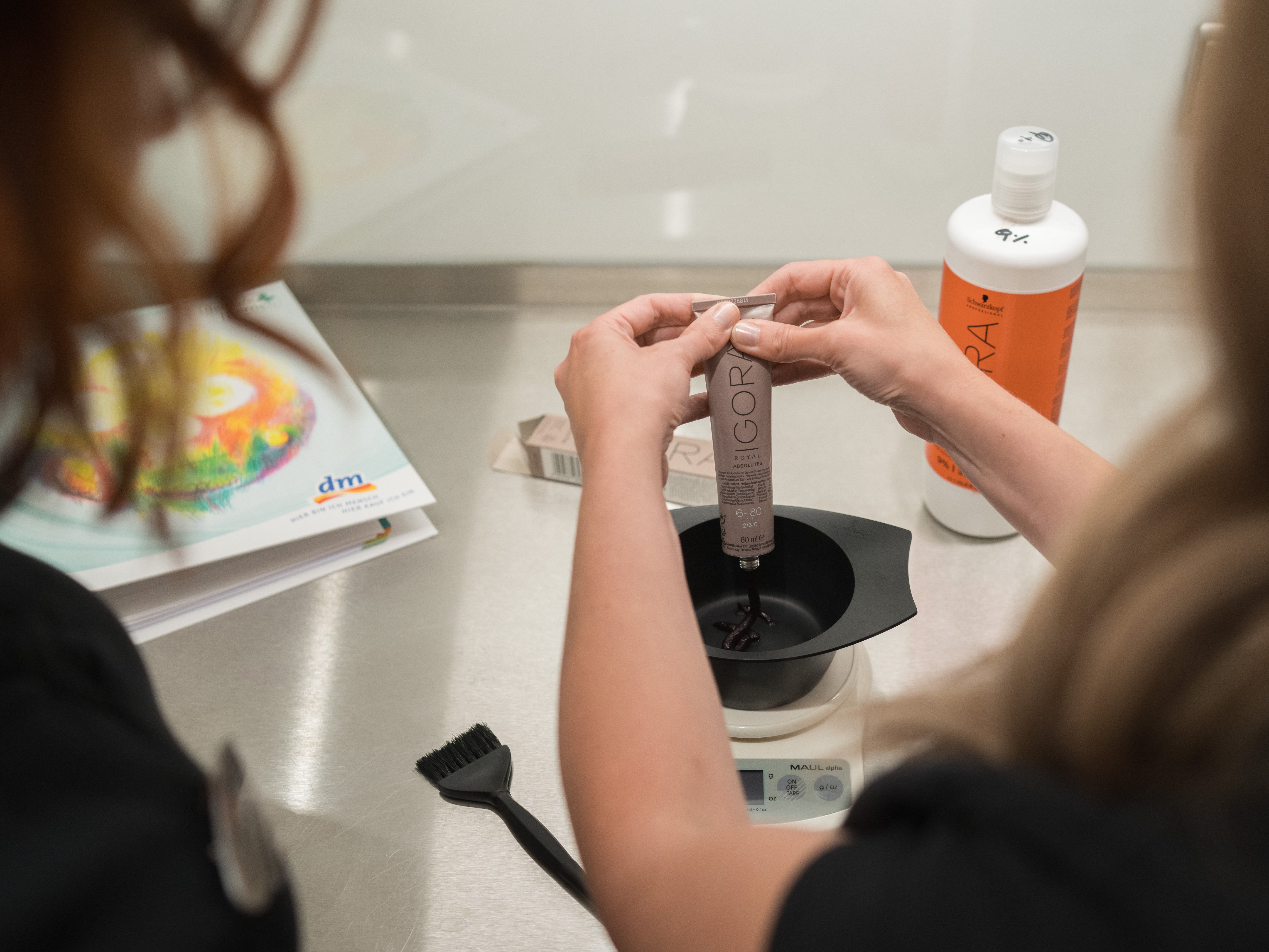 Eine Person hält eine Tube Haarfarbe über eine Schüssel und drückt den Inhalt hinein. Auf der Arbeitsfläche sind ein Pinsel, eine Waage und ein Haarpflegeprodukt sichtbar.