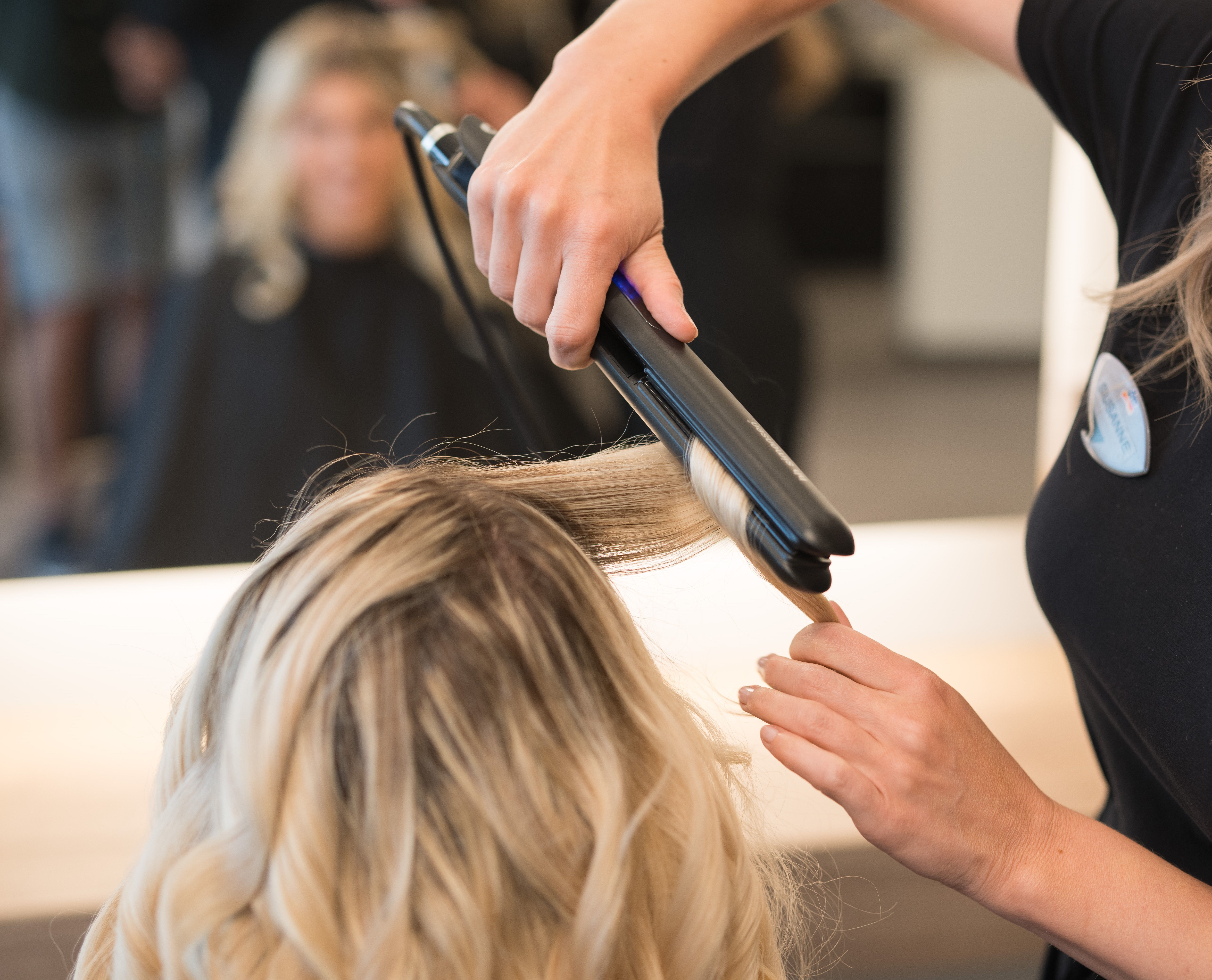 Ein Friseur benutzt ein Glätteisen, um das blonde Haar einer Frau in einem Salon zu locken, wobei ein Spiegel die Szene reflektiert.