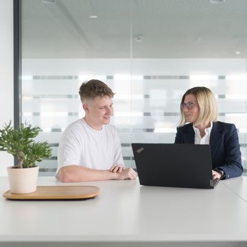 Eine Lehrling und eine Fachkraft sitzen an einem Tisch in einem modernen Besprechungsraum und unterhalten sich über Inhalte auf einem Laptop. Die Athmosphäre wirkt locker.