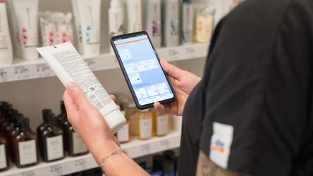 Eine Person steht vor einem Regal in einem dm Markt und hält ein Smartphone in der einen und ein Produkt in der anderen Hand. Auf dem Smartphone ist eine App geöffnet, die Informationen zu Produkten anzeigt.