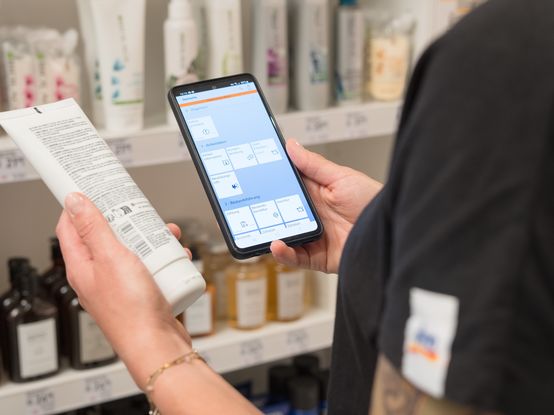 Eine Person steht vor einem Regal in einem dm Markt und hält ein Smartphone in der einen und ein Produkt in der anderen Hand. Auf dem Smartphone ist eine App geöffnet, die Informationen zu Produkten anzeigt.