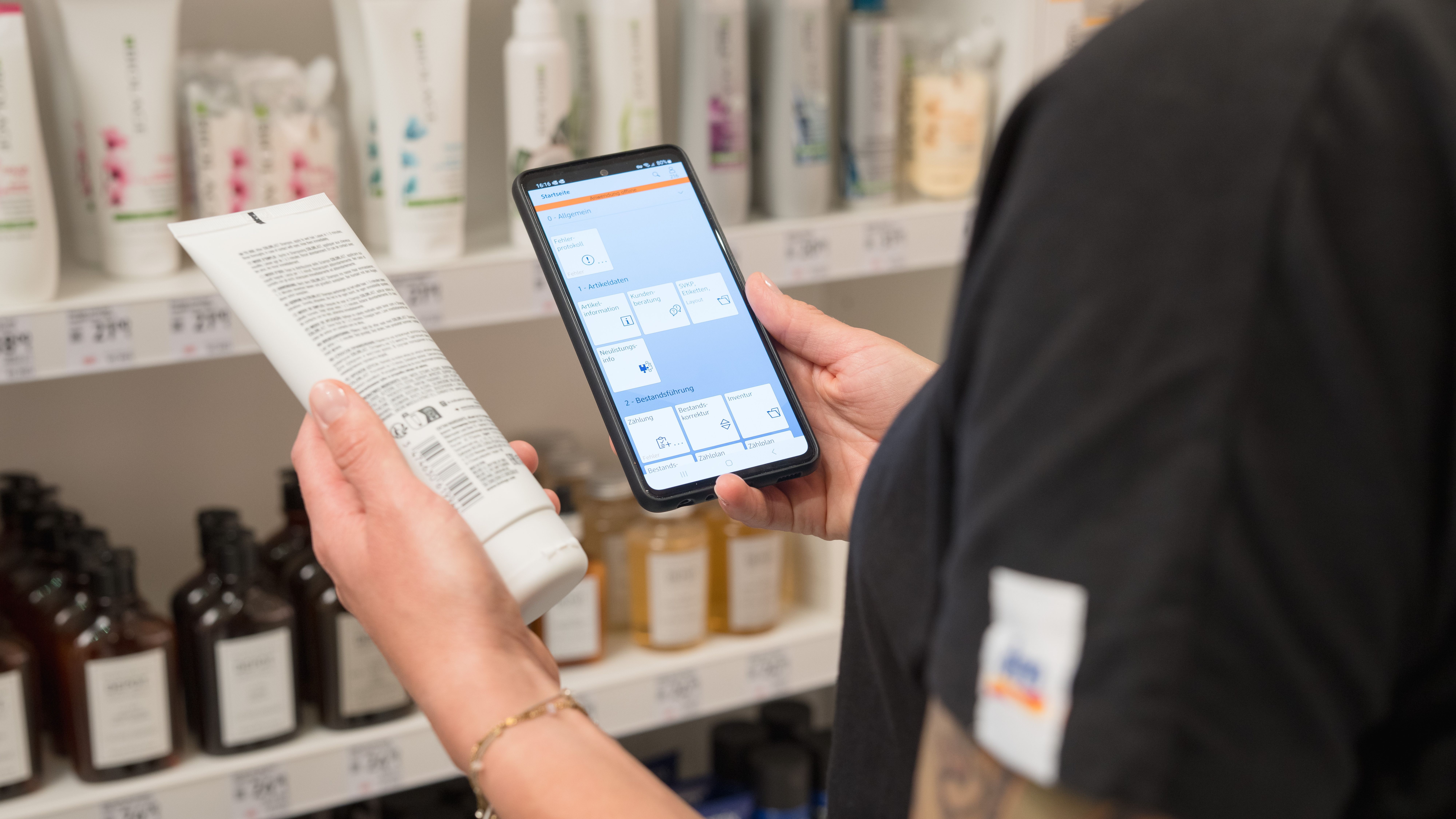 Eine Person steht vor einem Regal in einem dm Markt und hält ein Smartphone in der einen und ein Produkt in der anderen Hand. Auf dem Smartphone ist eine App geöffnet, die Informationen zu Produkten anzeigt.