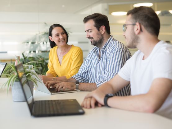 Eine Mitarbeiterin und zwei Mitarbeiter sitzen nebeneinander an der Highbench vor ihren Laptops und unterhalten sich miteinander.