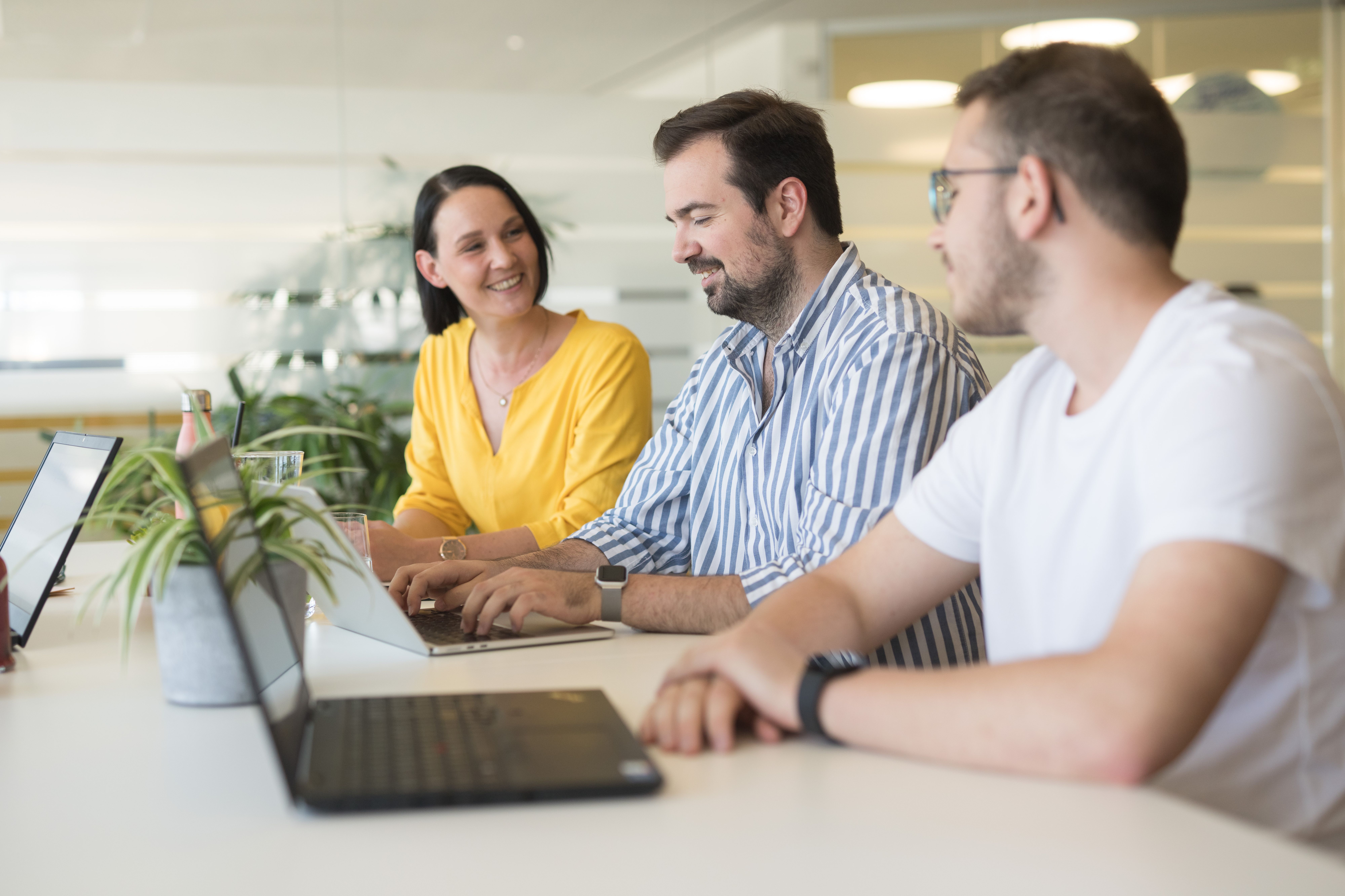 Eine Mitarbeiterin und zwei Mitarbeiter sitzen nebeneinander an der Highbench vor ihren Laptops und unterhalten sich miteinander.
