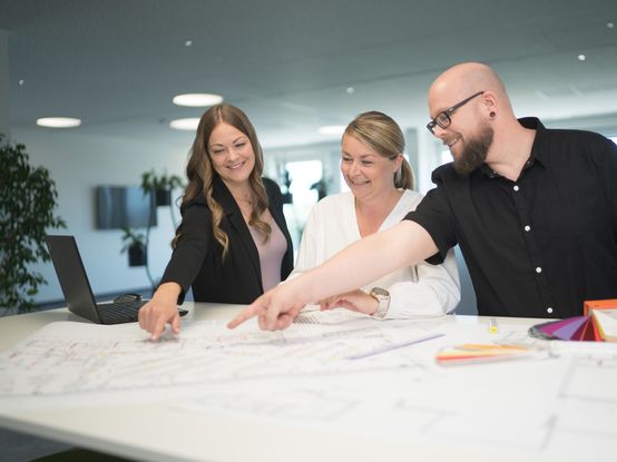 Zwei Mitarbeiterinnen und ein Mitarbeiter stehen an einer Highbench im Büro und betrachten gemeinsam Filialpläne. Der Mitarbeiter auf der linken Seite und die Mitarbeiterin auf der rechten Seite zeigen auf einen bestimmten Bereich eines Plans und lächeln.