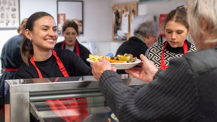 Eine Frau reicht einem Mann en Teller mit Essen bei einer Essensausgabe. Im Hintergrund sind noch weitere Personen zu sehen.